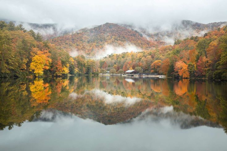 a lake surrounded by trees covered in fall foliage and foggy skies with mountains in the background