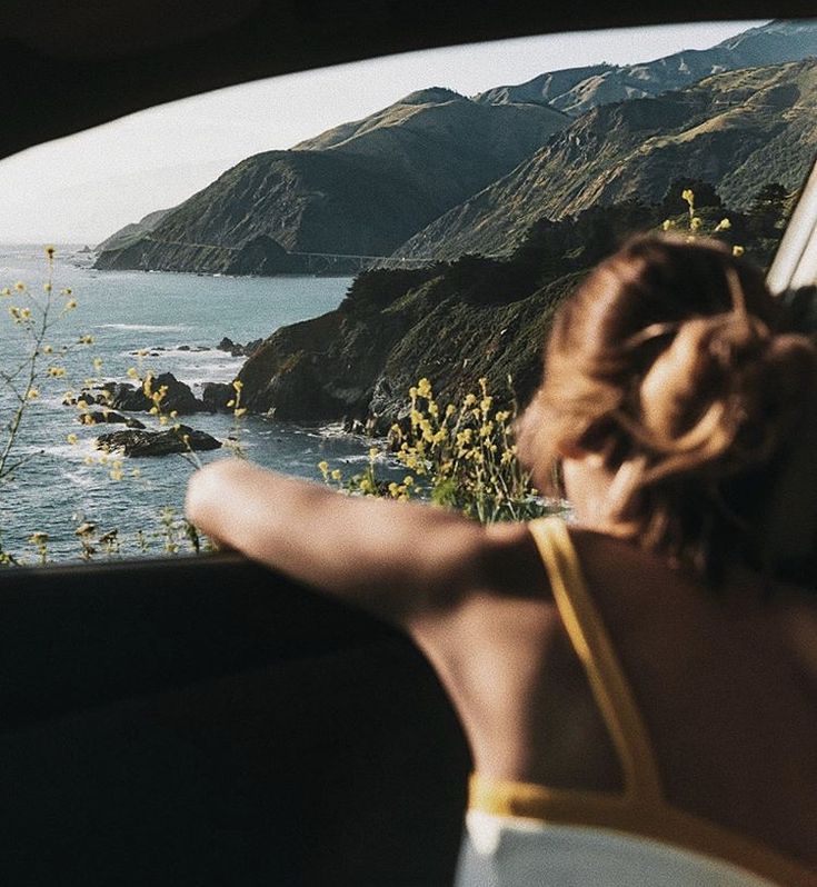 a woman is looking out the window of a car at the ocean and mountains in the distance