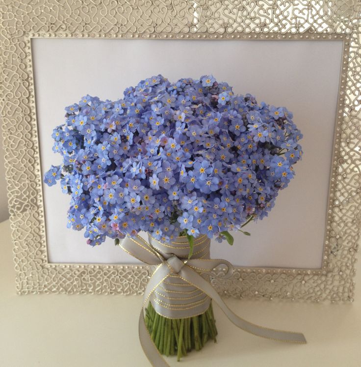 a bouquet of blue flowers in a vase with a ribbon tied around the bottom and an ornate frame behind it