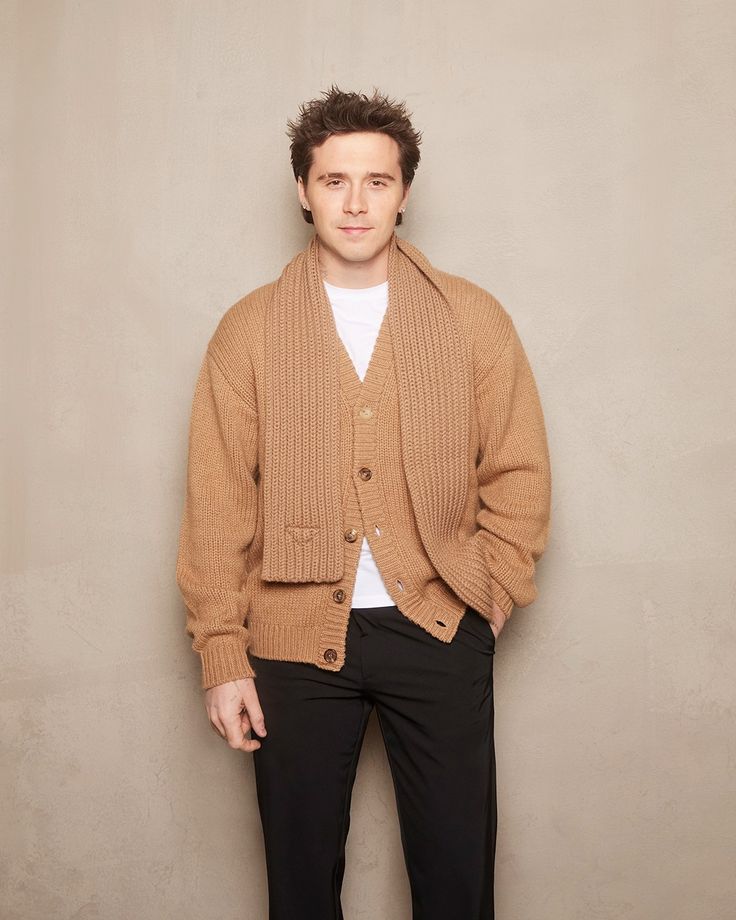 a man standing in front of a wall wearing a brown cardigan sweater and black pants