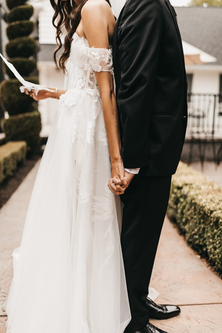 a bride and groom standing next to each other in front of a house holding hands