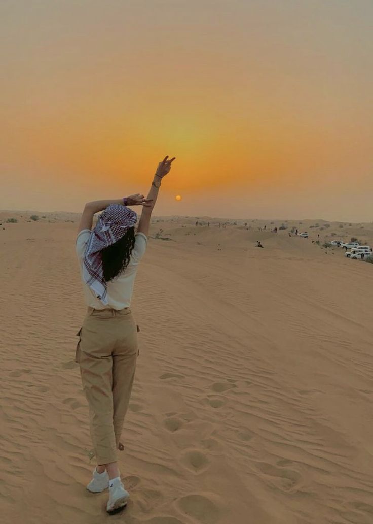 a woman standing in the sand with her arms up to the sun setting behind her