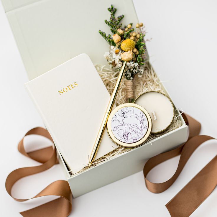 an open box containing candles, flowers and a note book with ribbon around it on a white surface