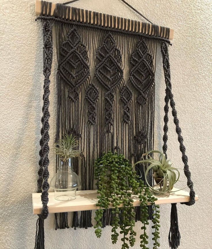 some plants are sitting on a shelf in front of a wall hanging with macrami