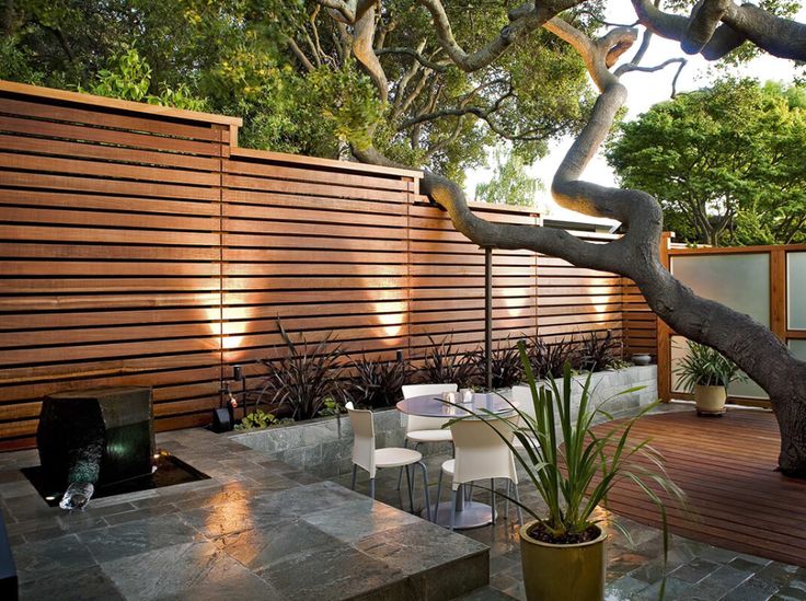 an outdoor dining area with wooden fence and white chairs in the foreground, surrounded by greenery