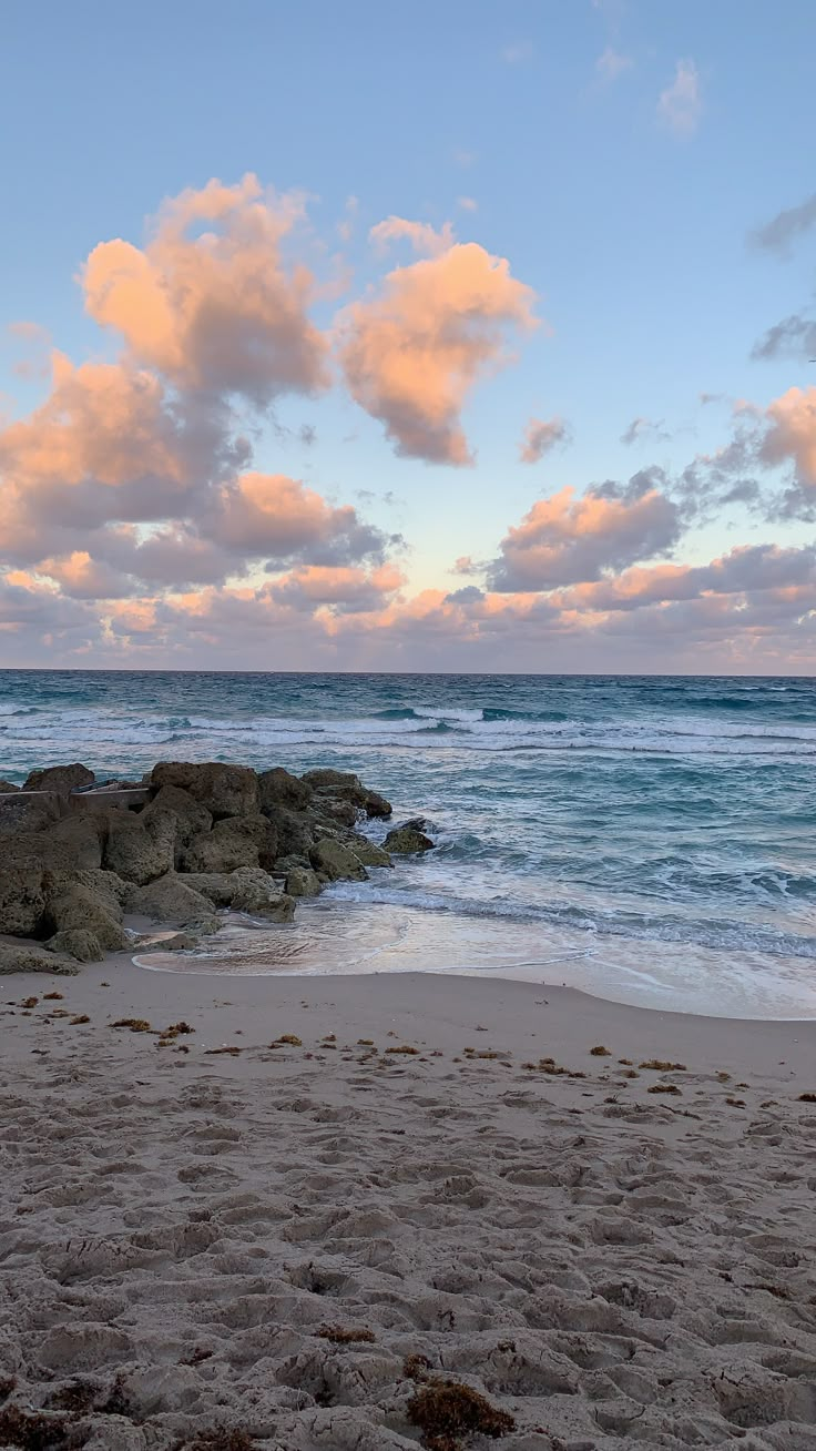 the beach has waves coming in from the ocean and rocks on the shore, under a cloudy blue sky with white clouds