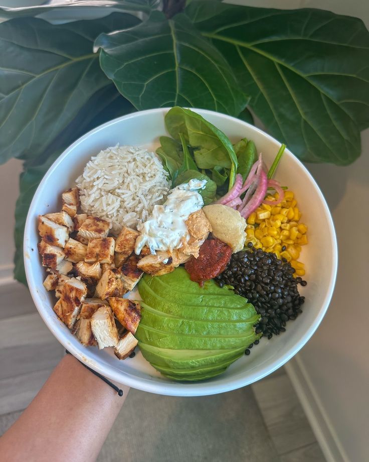 a person holding a white bowl filled with different types of food and greens on top of it