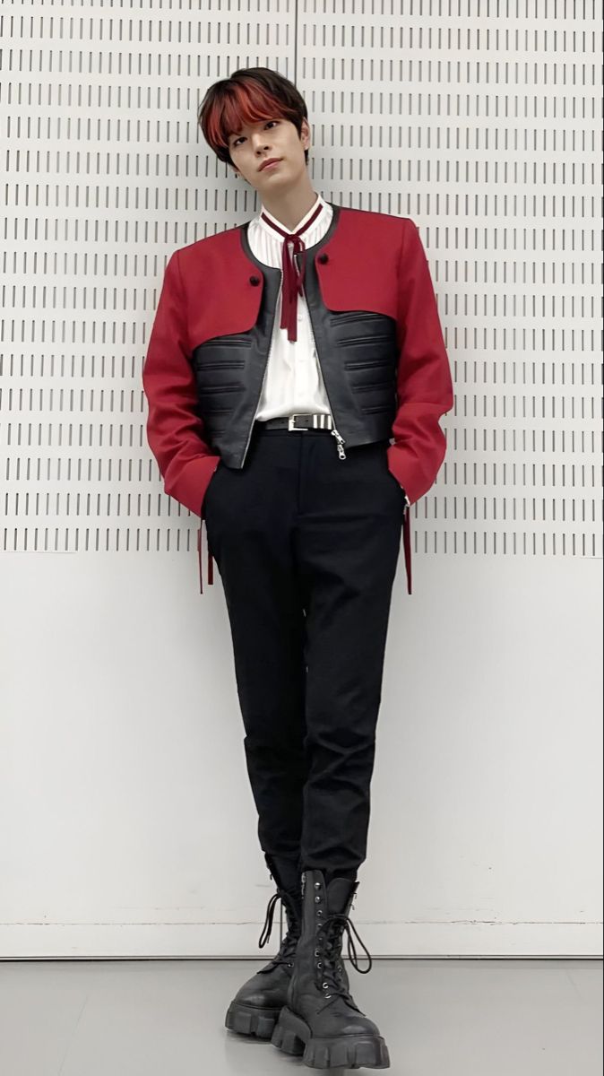 a man in a red jacket and black pants stands against a wall with his hands on his hips