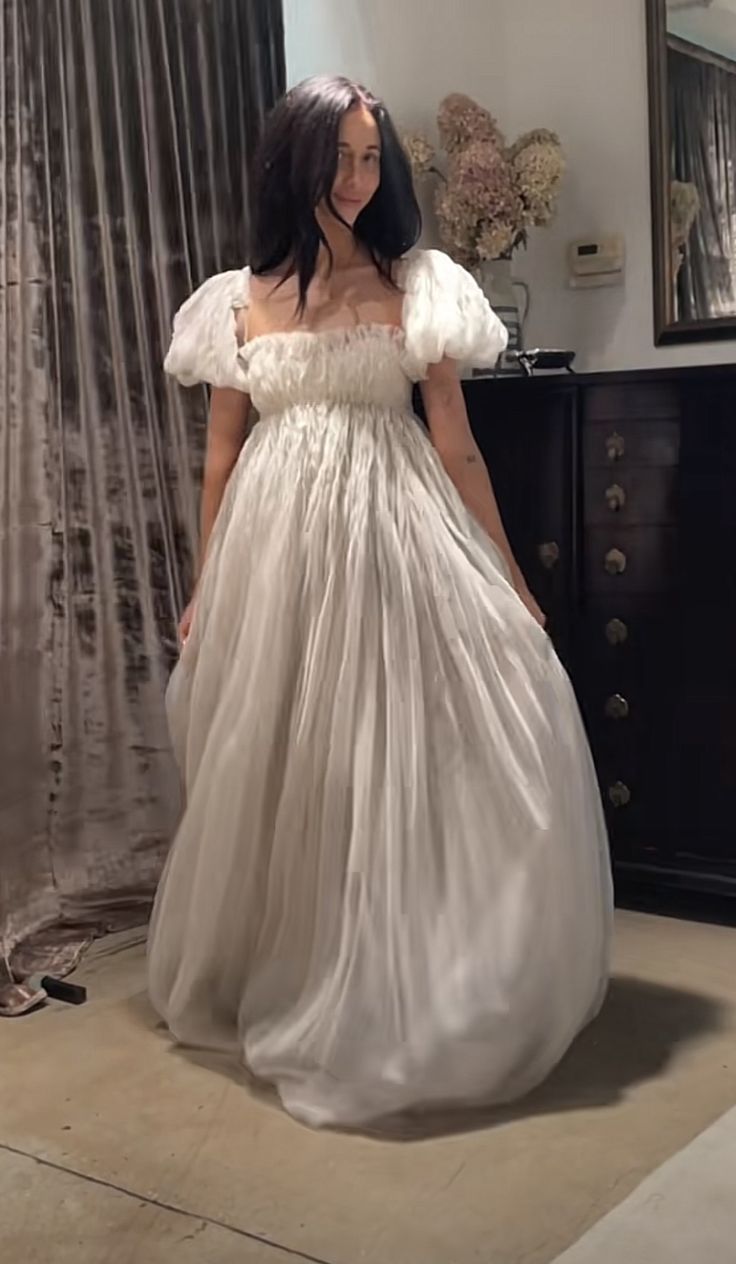 a woman wearing a white dress standing in front of a dresser and looking at the camera