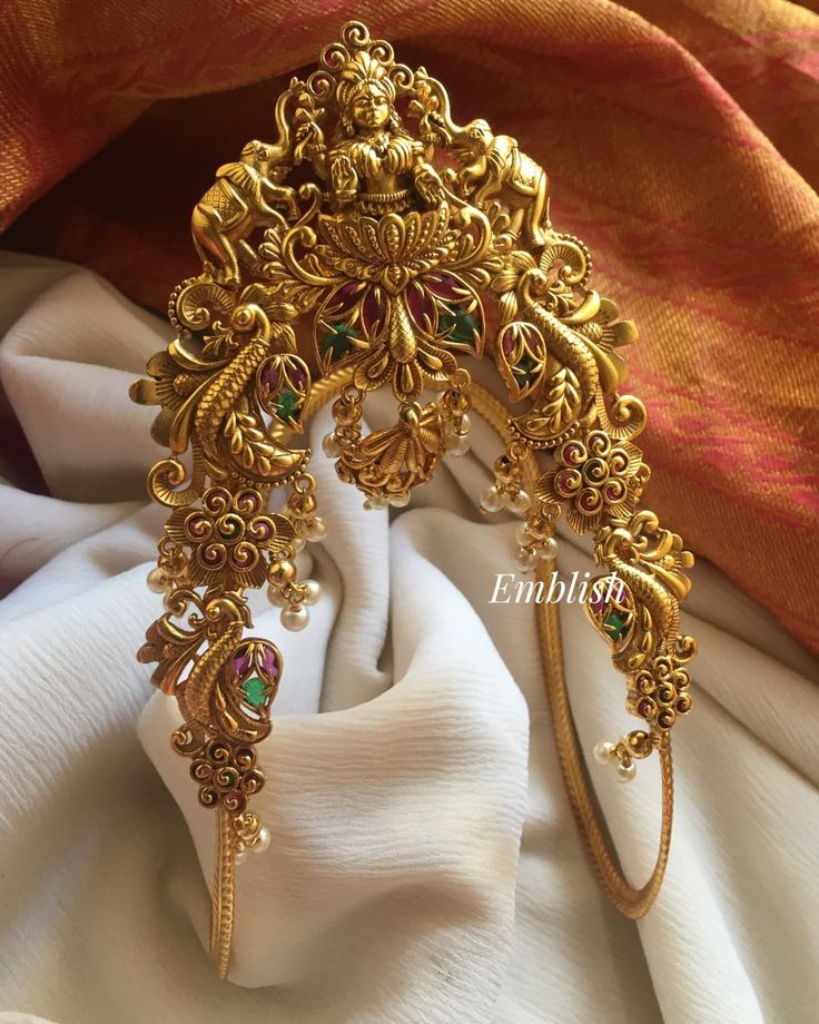 an ornate gold head piece with green and red stones on it's side, sitting on a white cloth