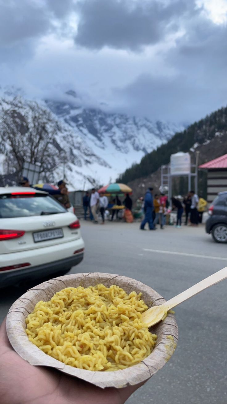 a hand holding a paper bowl filled with food and a wooden spoon in front of mountains