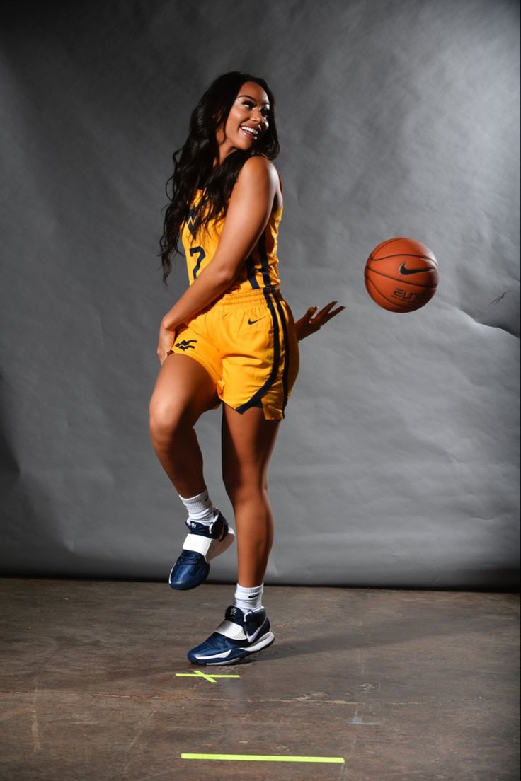 a woman in a yellow uniform holding a basketball and posing for a photo on a gray background