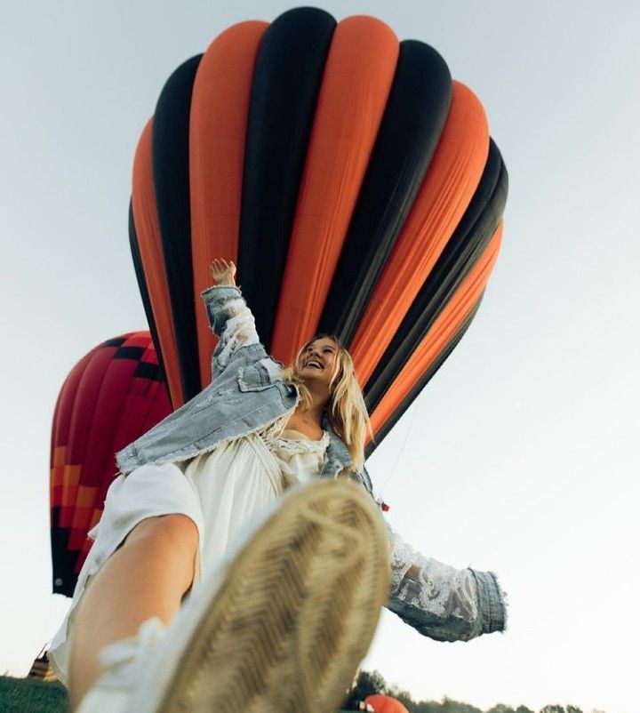 a woman in white shirt and jeans standing next to hot air balloon with her feet up