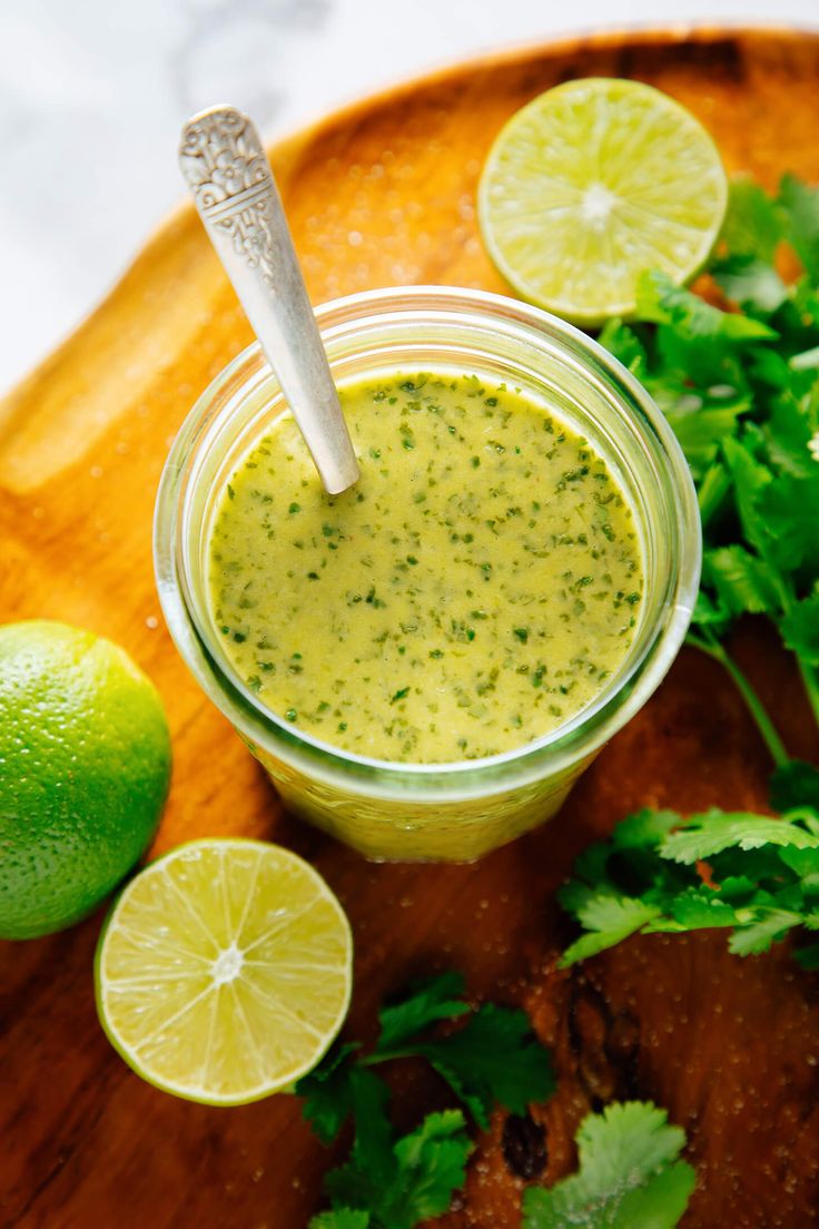 a small jar filled with green sauce next to limes and cilantro on a cutting board