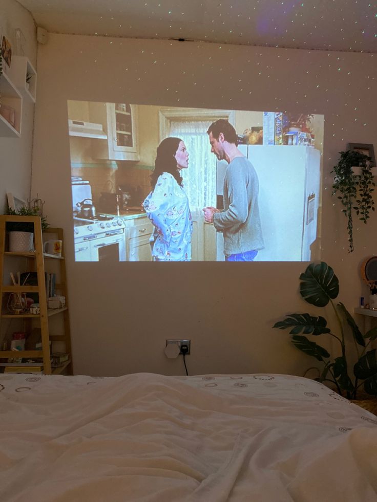 a man and woman standing in front of a screen on the wall next to a bed