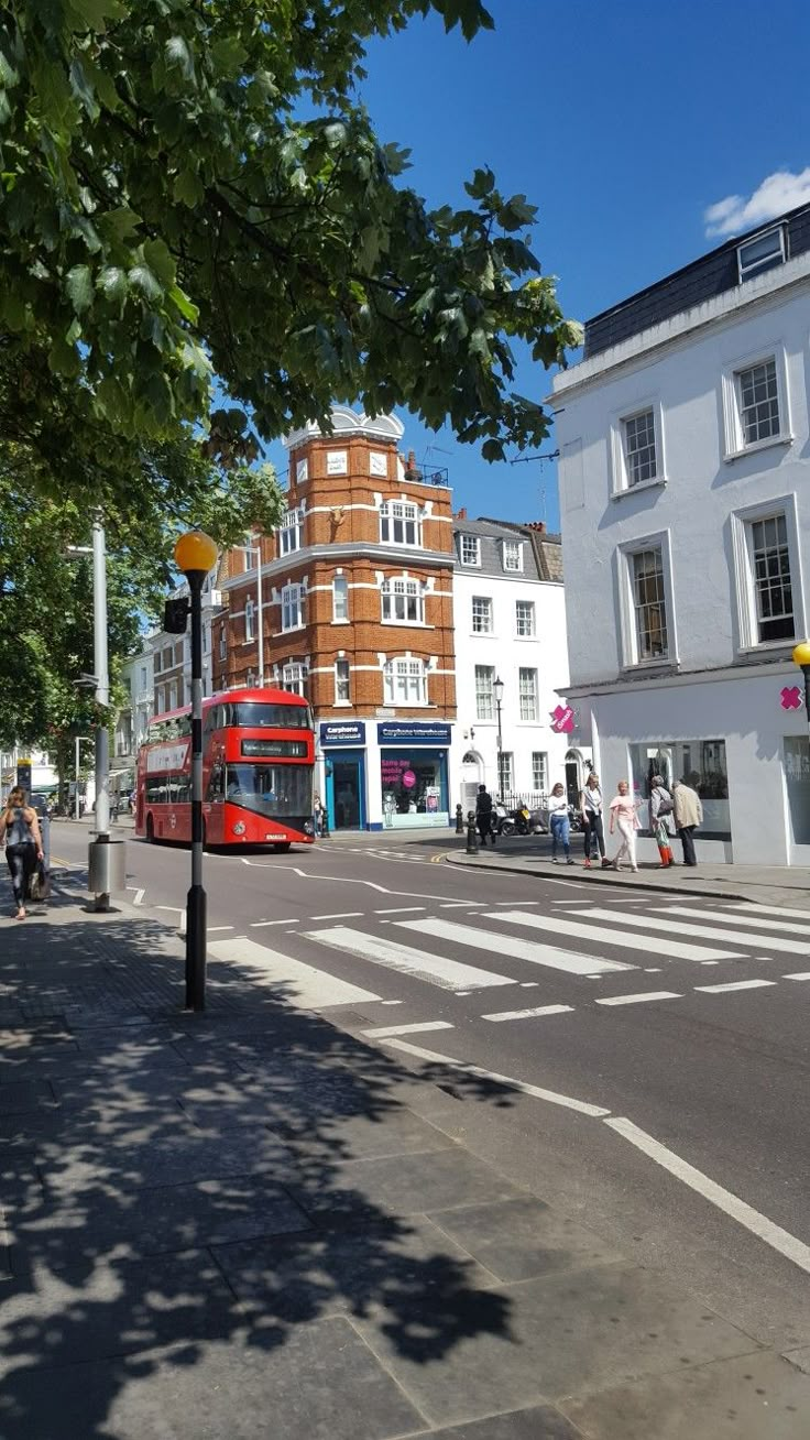 a red double decker bus driving down a street next to tall buildings and people walking on the sidewalk