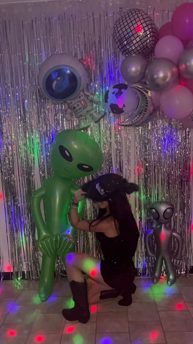 a woman kneeling down next to an alien balloon in front of balloons and streamers