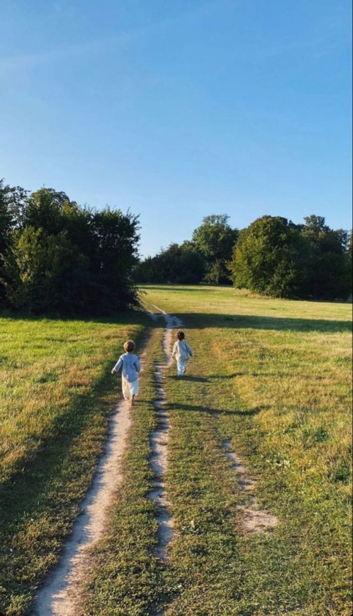 two people walking down a dirt road in the middle of a field