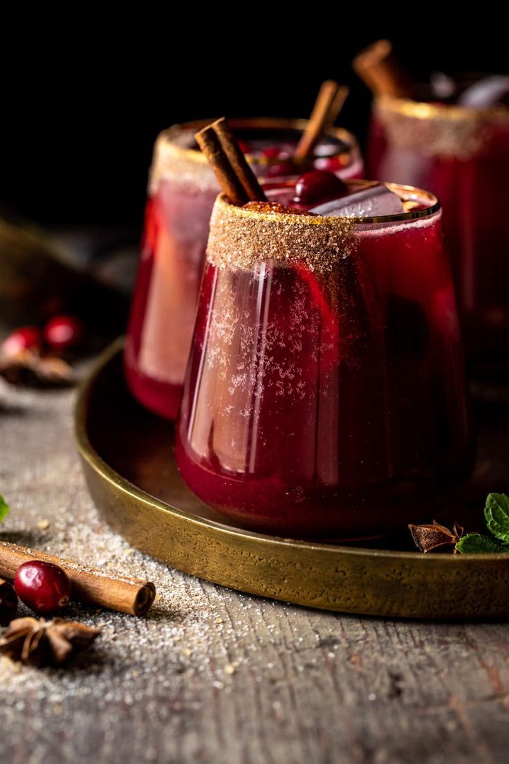 two glasses filled with red liquid and cinnamon sticks on top of a wooden tray next to spices