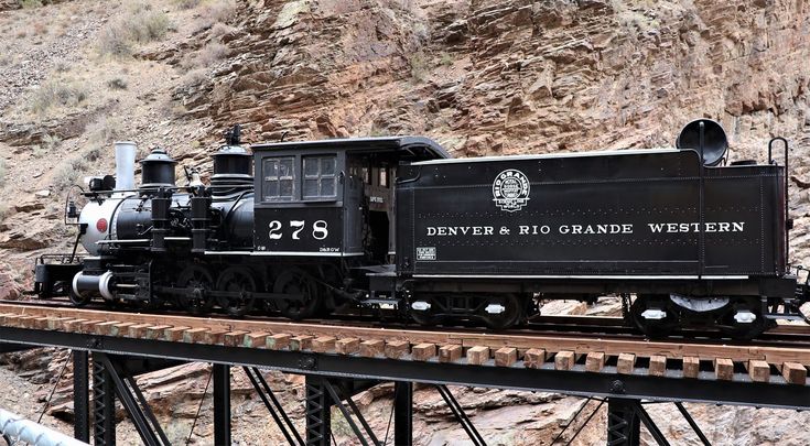 an old fashioned train is on the tracks in front of a rocky mountain side area