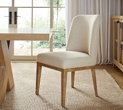 a white chair sitting in front of a window next to a wooden table and dresser