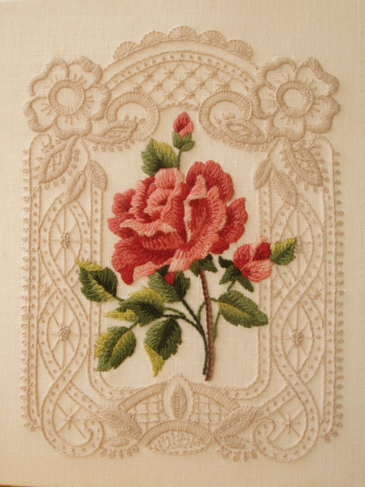 an embroidered rose with green leaves and pink flowers on a white linen background, framed by intricate lace