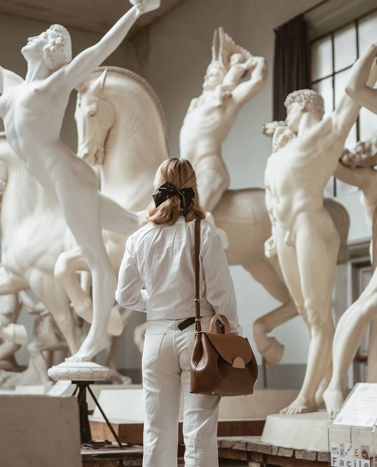 a woman standing in front of some white statues and holding a brown purse with her right hand