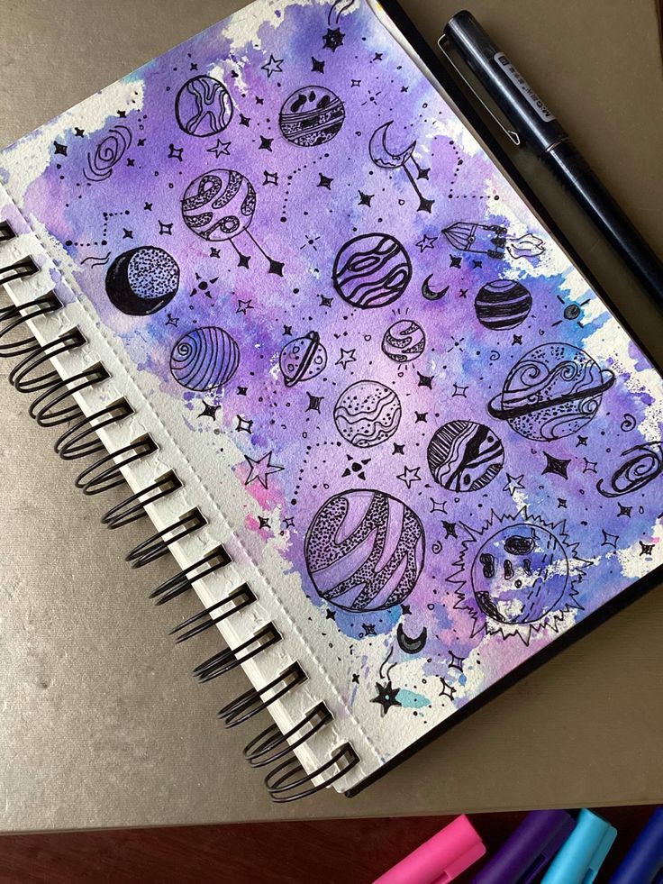 an open notebook with space drawings on it next to colored markers and pencils in the background