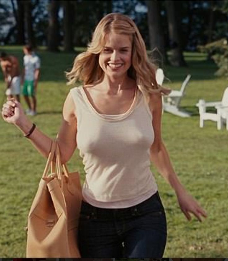 a woman walking across a lush green field holding a brown bag and smiling at the camera