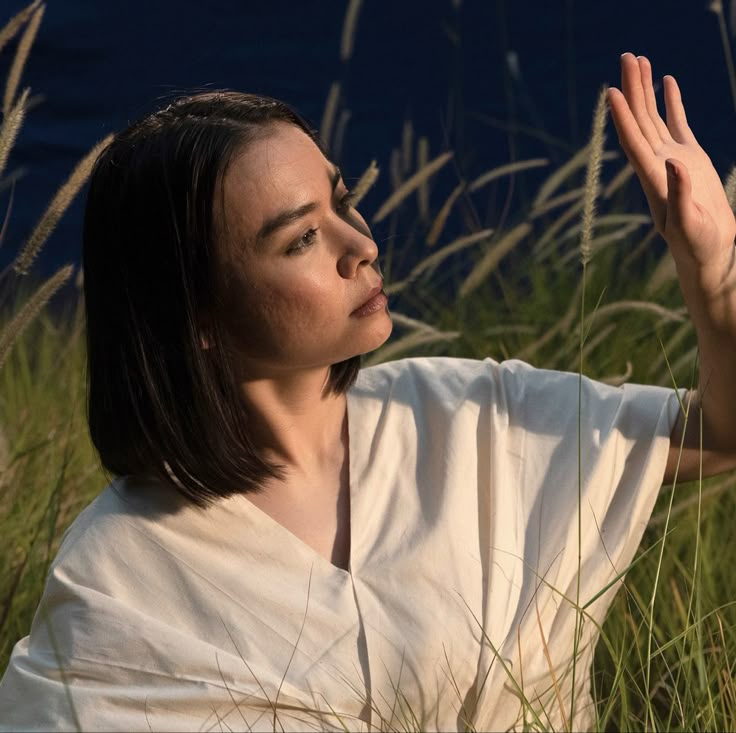 a woman sitting in tall grass holding her hand up to the side with both hands