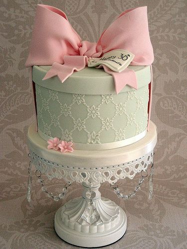 a three tiered cake decorated with pink bows and lace on a white pedestal against a damask wall