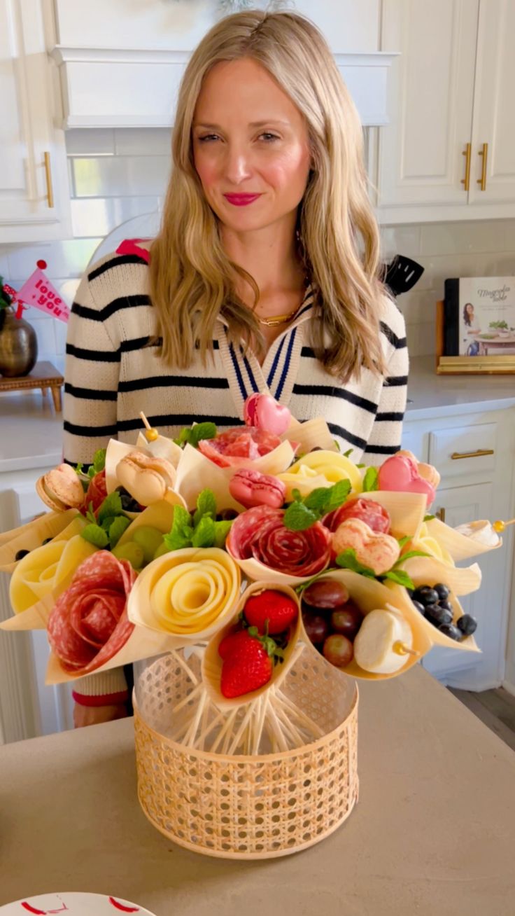 a woman standing in front of a table holding a vase filled with fruit and flowers