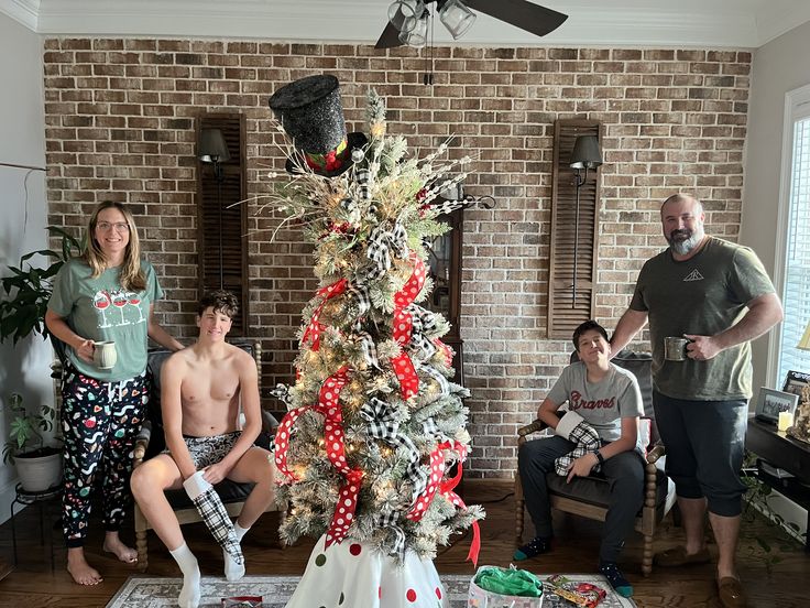 a group of people standing around a christmas tree in front of a brick wall and ceiling fan