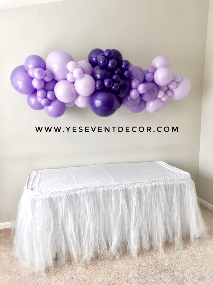 the balloon wall is decorated with purple and lavender balloons, while white tulle skirts are below it