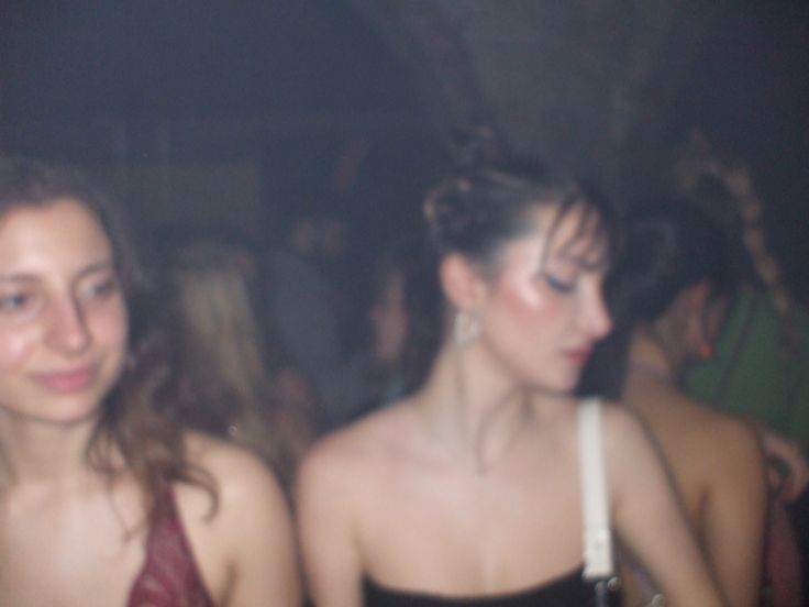 two young women standing next to each other in a room full of people at a party