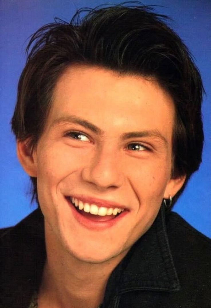 a man with black hair smiling and wearing a necklace on it's neck, in front of a blue background