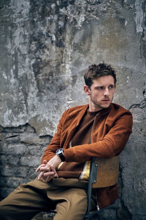 a man sitting on top of a chair next to a stone wall and wearing a brown jacket