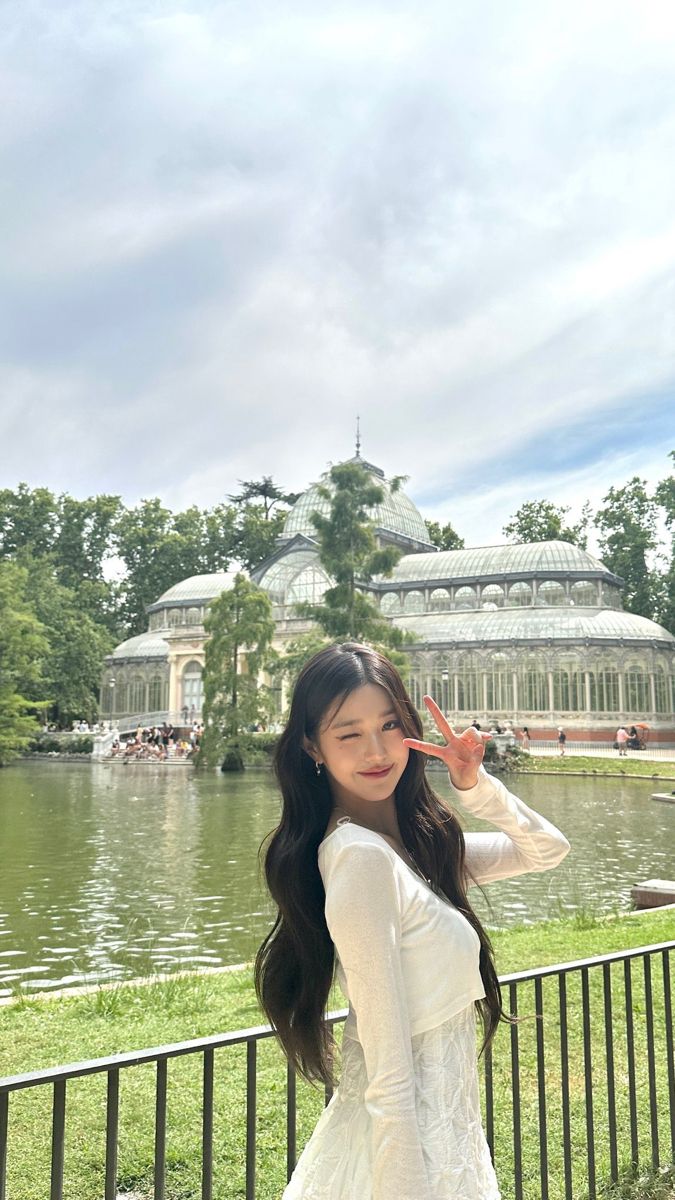 a woman in a white dress is posing for the camera near a pond and glass house