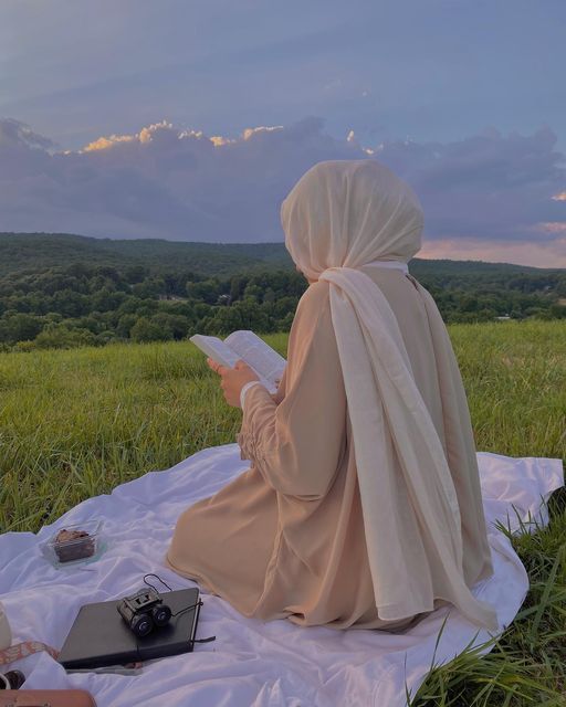 a woman sitting on top of a blanket in a field holding a book and looking at the sky