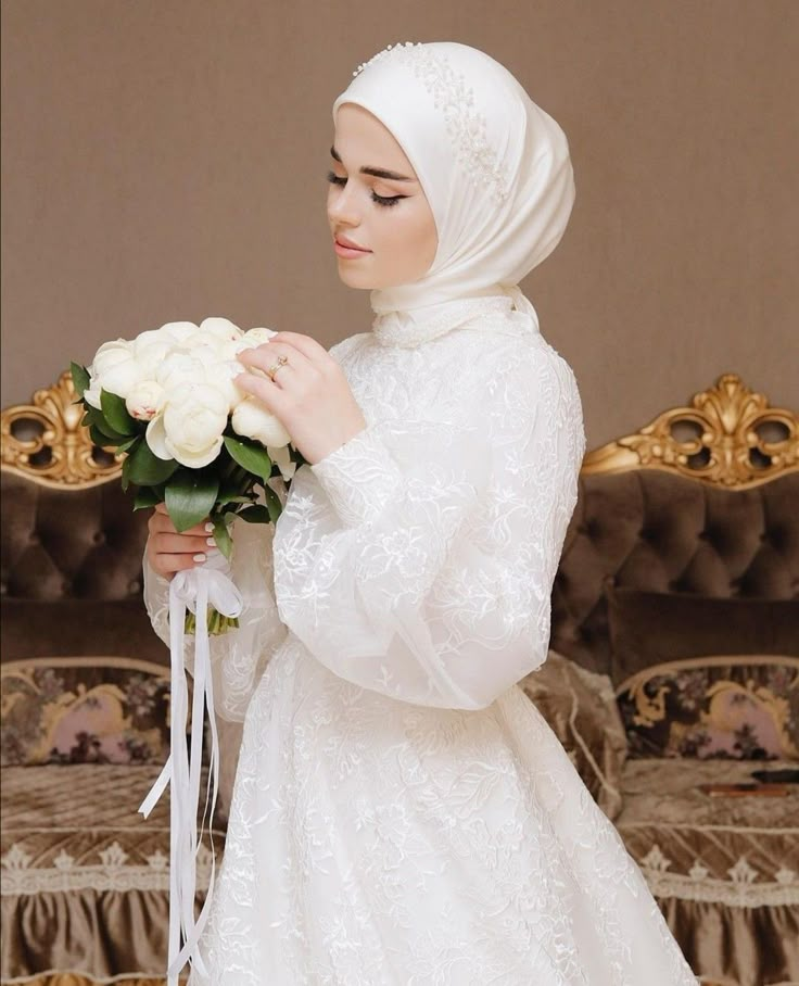 a woman in a white dress holding a bouquet of flowers and wearing a headscarf