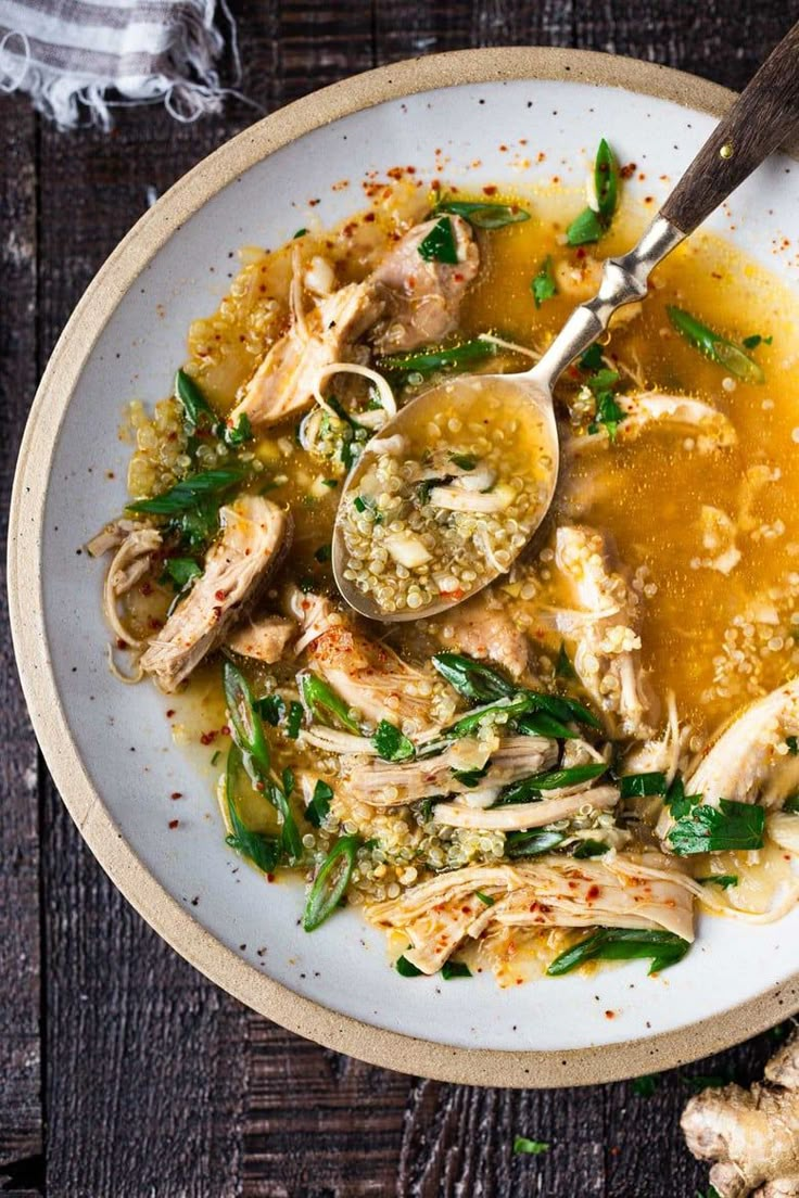 a white bowl filled with chicken and vegetable soup on top of a wooden table next to two spoons