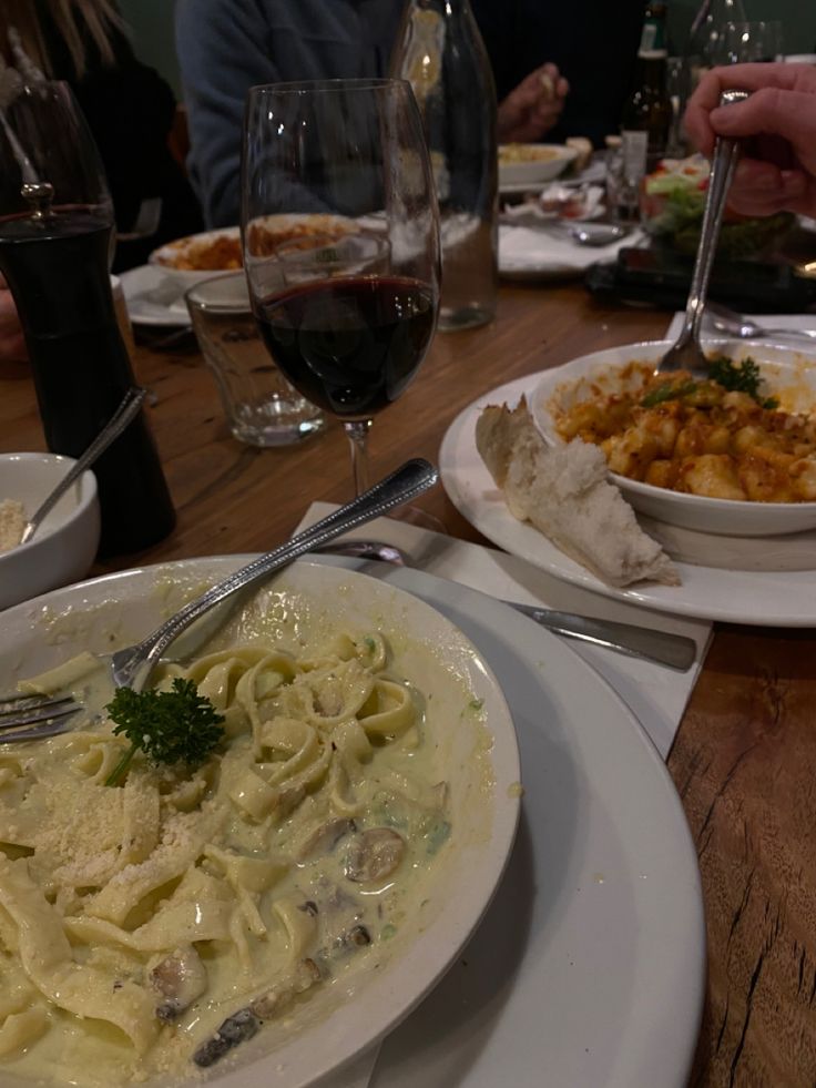 two plates of pasta and wine on a wooden table with people sitting at the table