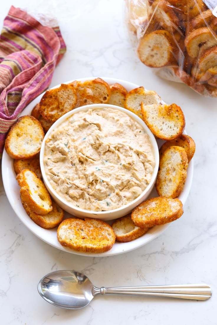 a white bowl filled with dip surrounded by toasted bread on a marble counter top