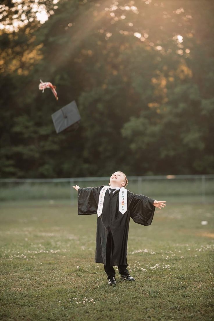 a young boy in graduation gown standing on grass with his arms out to the sky