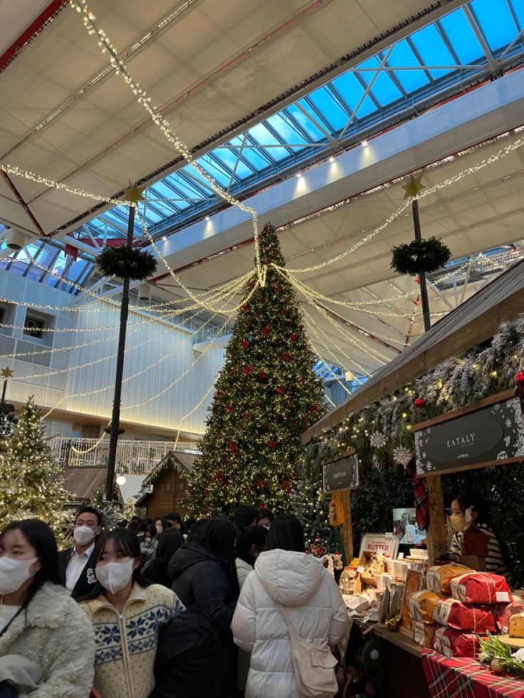 people wearing face masks are shopping at a christmas tree in a mall with lights and decorations on the ceiling