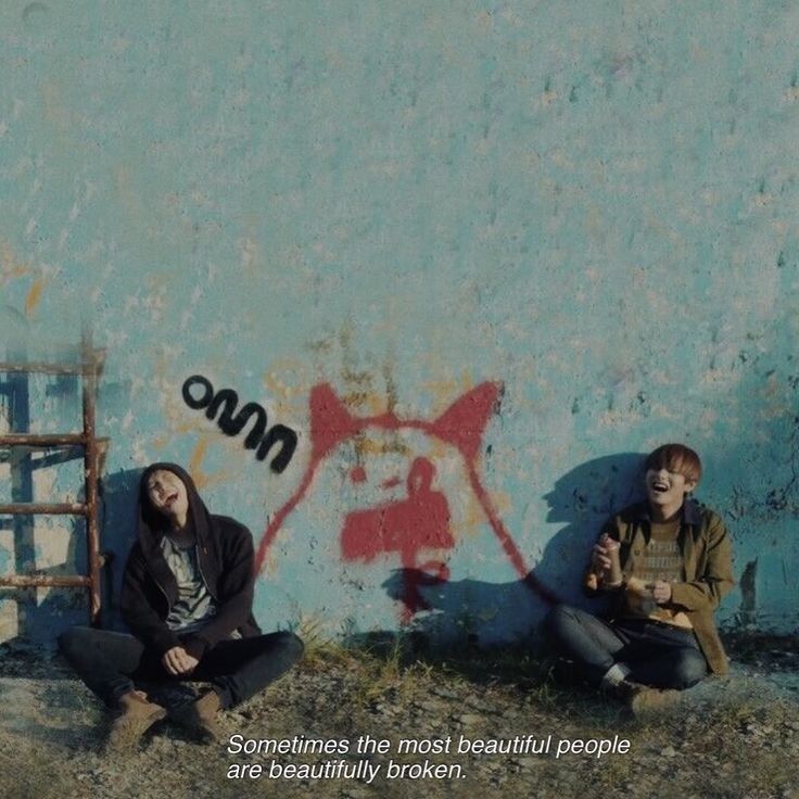 two people sitting on the ground next to a wall with graffiti and words written on it