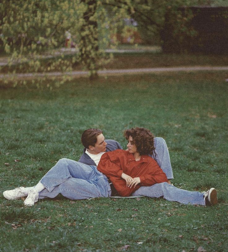 two people laying on the grass in front of a tree and one person holding another