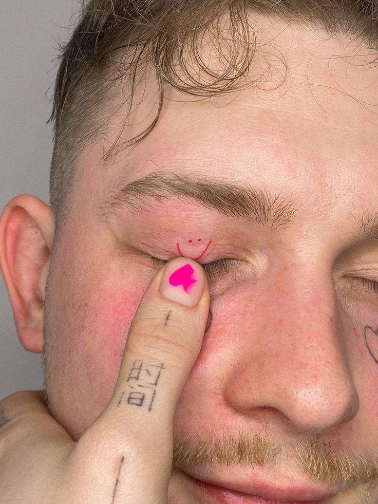 a man with a pink heart tattoo on his left eye and hand next to his face