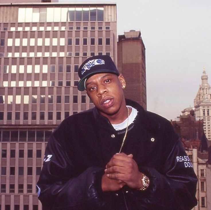 a man standing in front of tall buildings wearing a black jacket and hat with his arms crossed