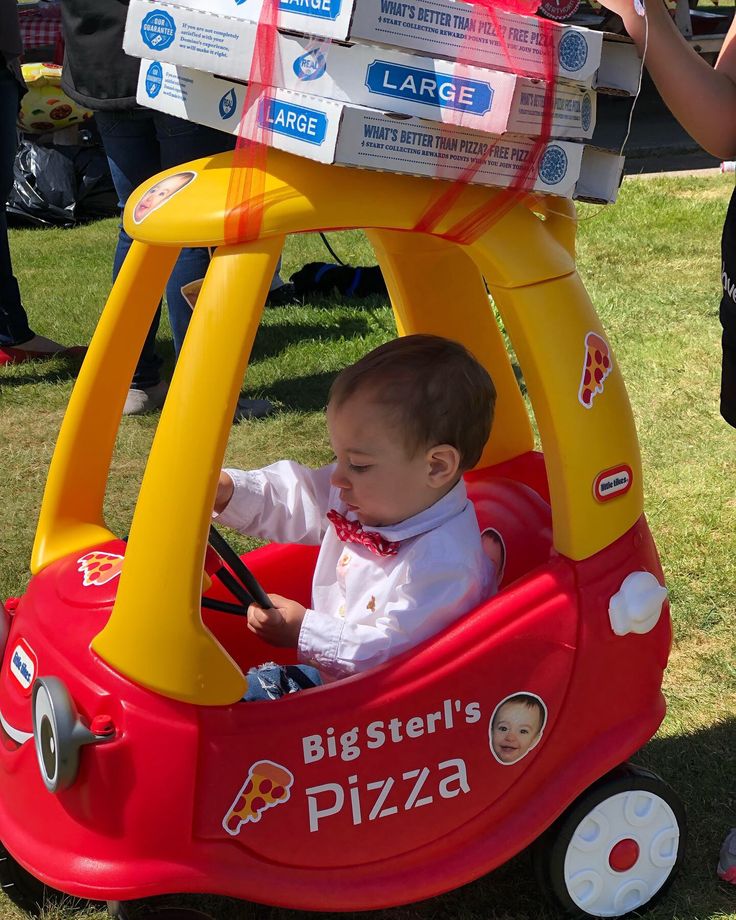 a baby sitting in a red and yellow toy car with pizza boxes on it's roof